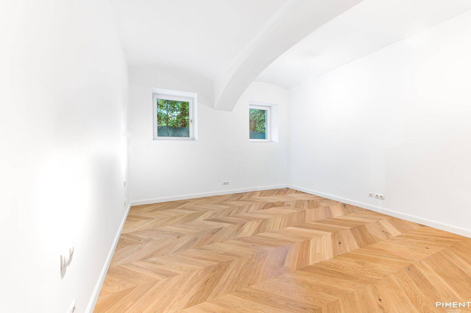 Helles Schlafzimmer mit Fischgrätparkett und zwei Fenstern, die viel Tageslicht hereinlassen.