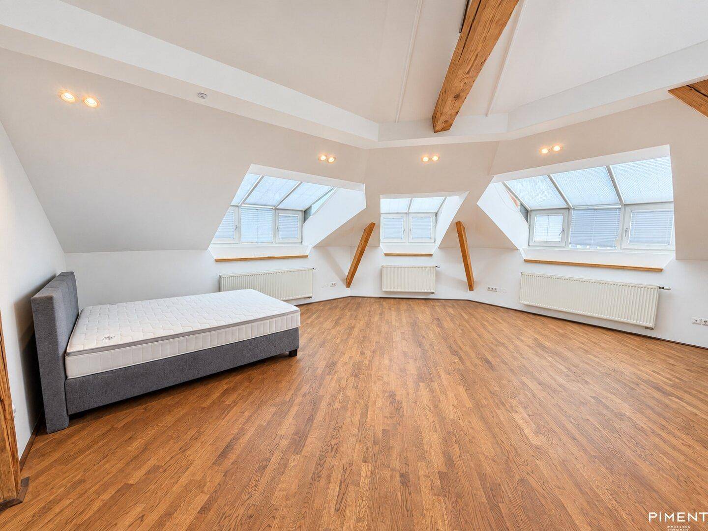 Helles Schlafzimmer im Dachgeschoss mit drei Dachfenstern, Holzbalken und einem Bett.