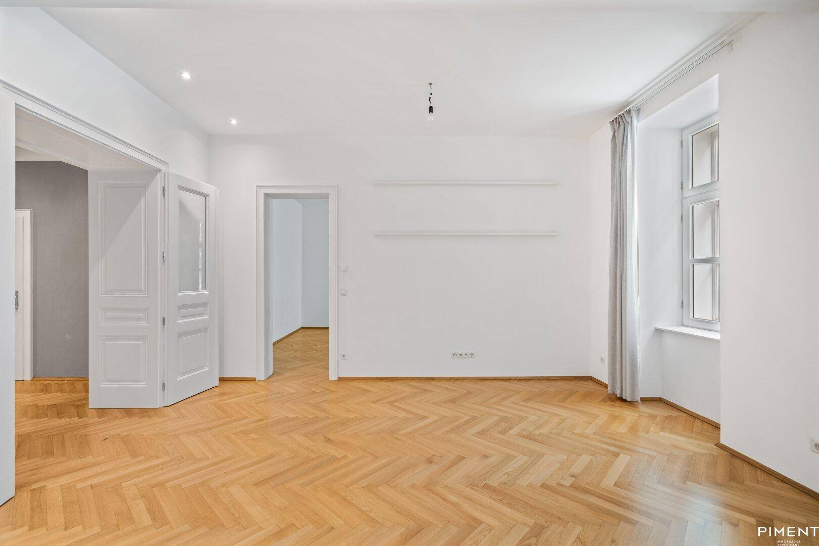 Geräumiges Wohnzimmer mit Fischgrätparkett, hohen Decken und großen Fenstern.