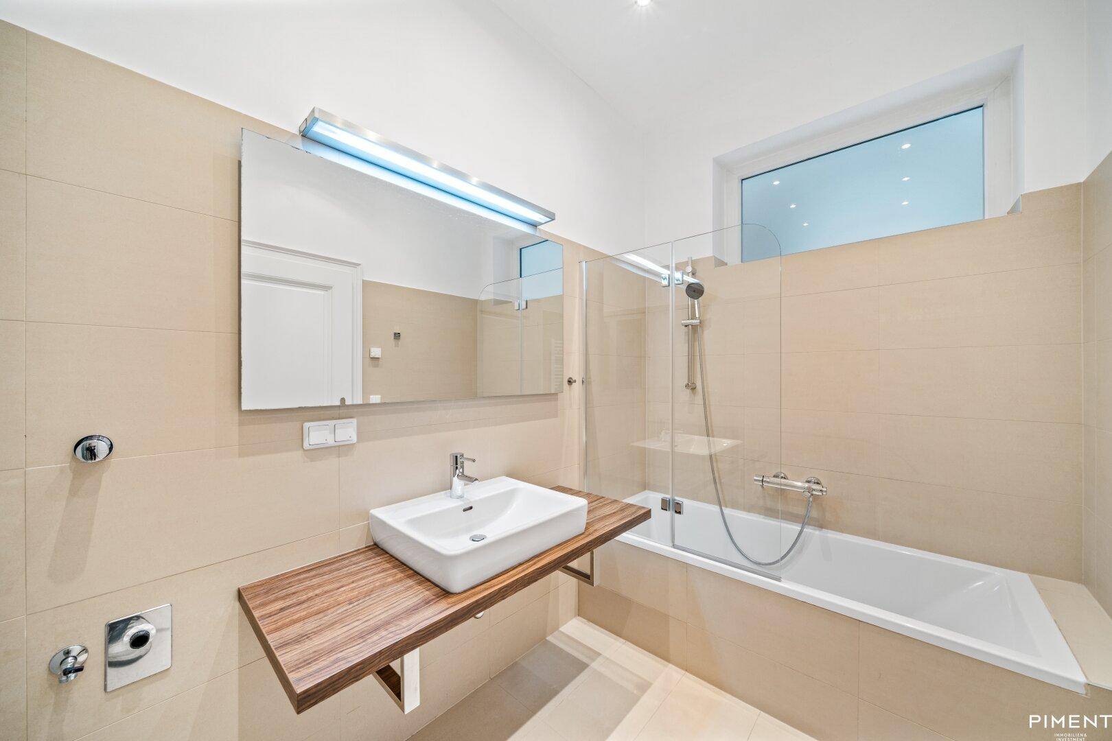 Helles Badezimmer mit Badewanne, Duschabtrennung, Fenster und modernem Waschtisch.