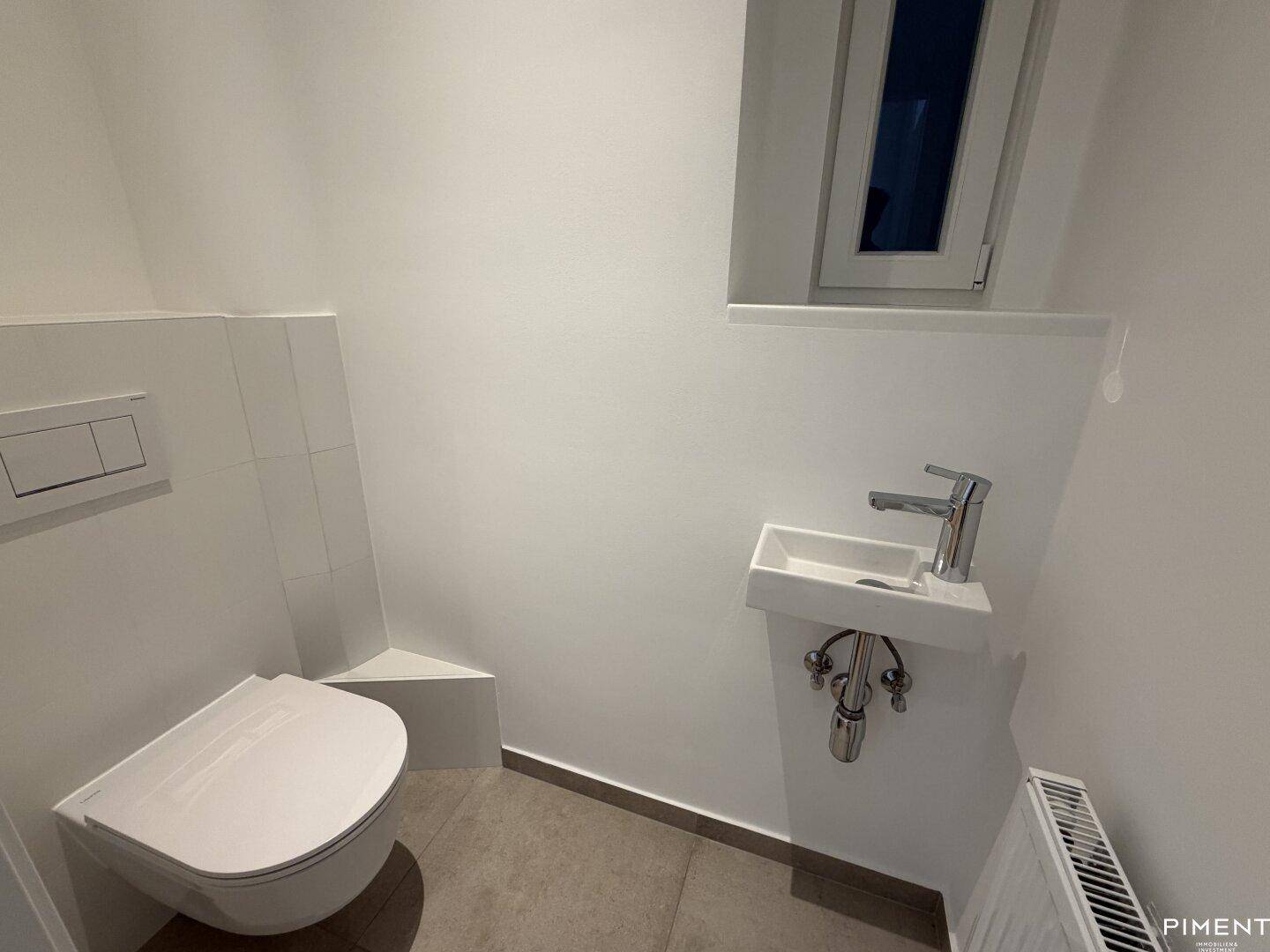 Separates WC mit wandhängender Toilette, kleinem Waschbecken und einem Fenster für Tageslicht.