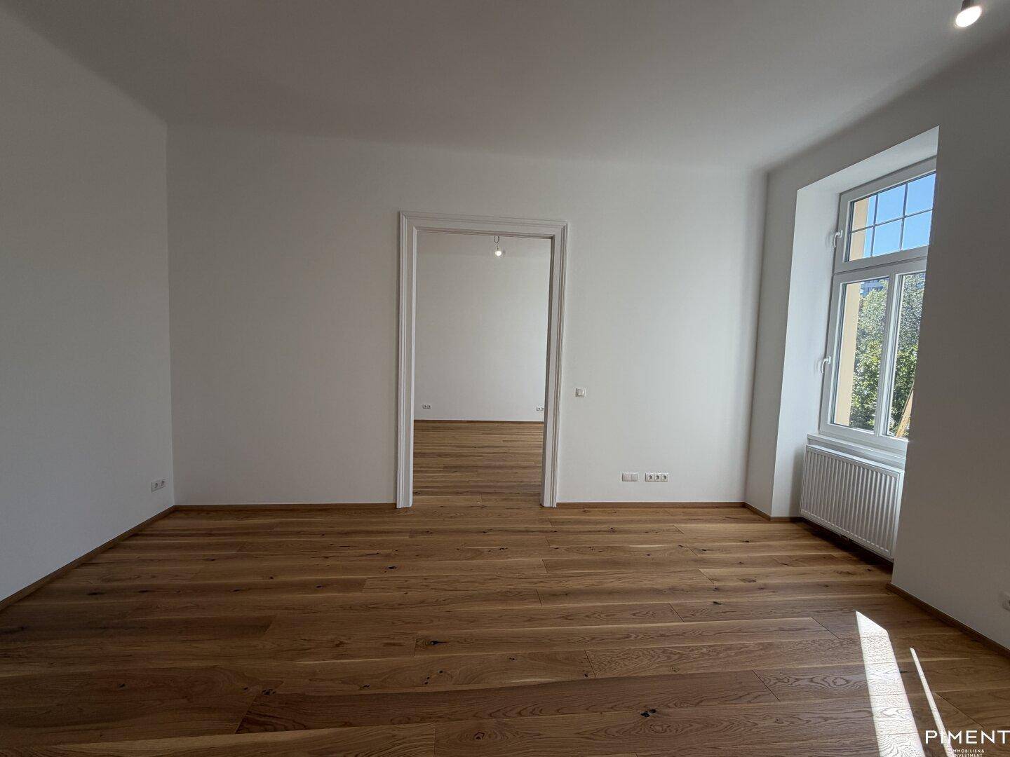 Geräumiges Zimmer mit hellem Parkettboden und einem Fenster, das den Blick ins Freie freigibt.