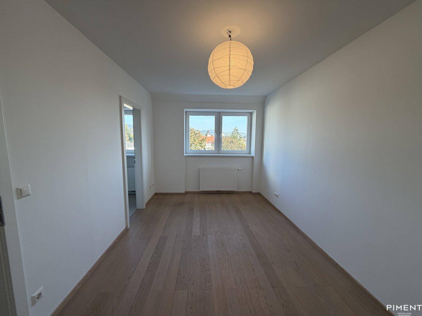 Helles Zimmer mit Parkettboden und einem Fenster, das den Blick ins Grüne freigibt.