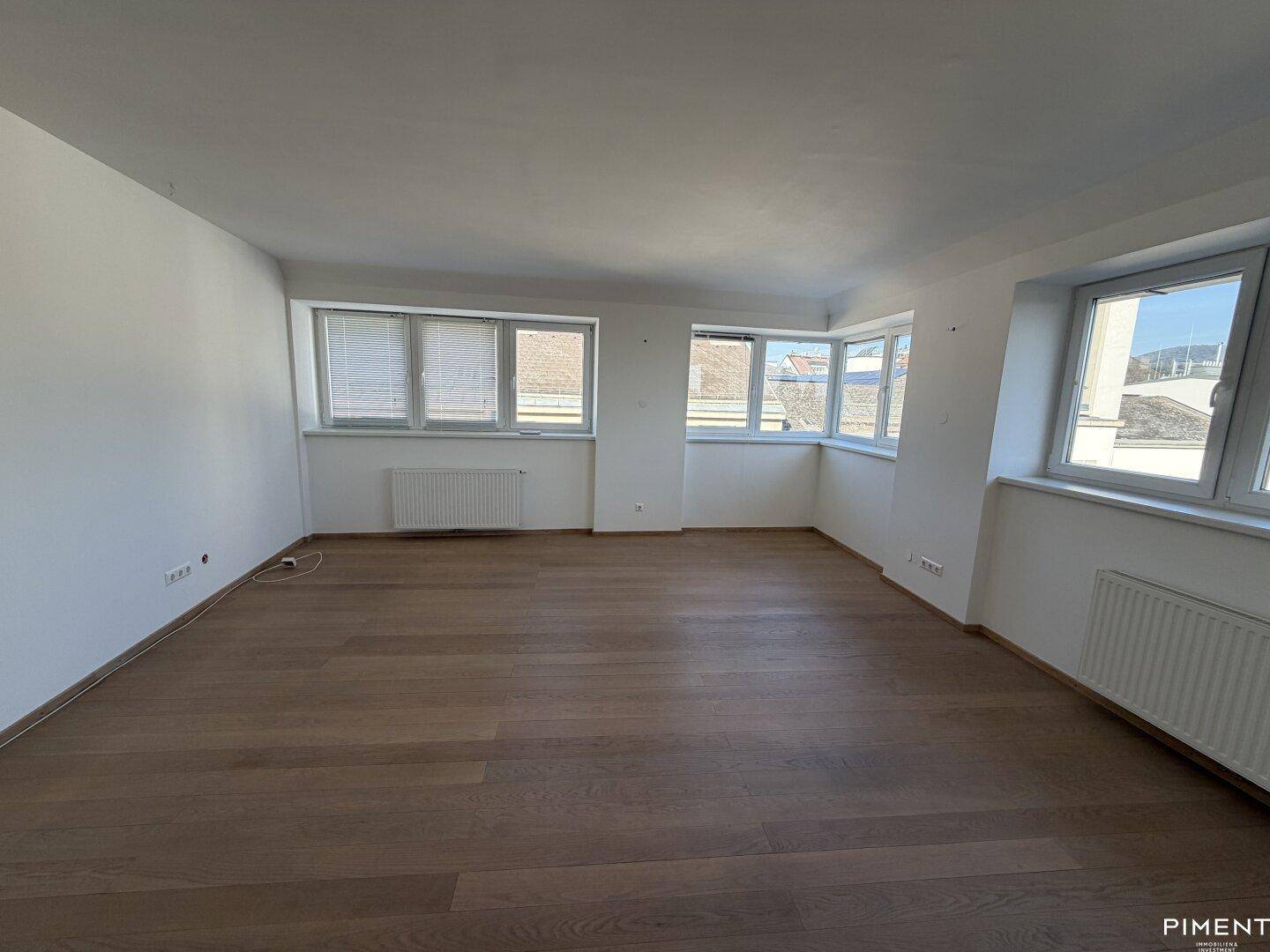Geräumiges Wohnzimmer mit Holzboden und mehreren Fenstern, die eine helle Atmosphäre schaffen.