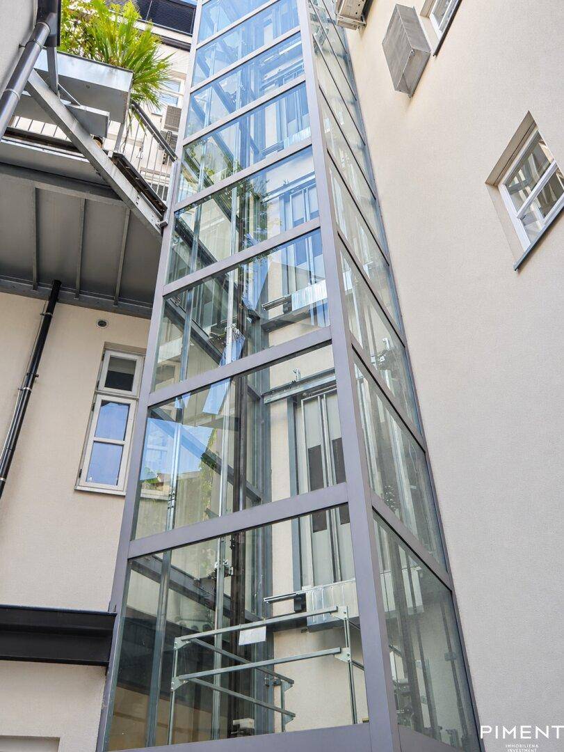 Außenansicht eines modernen Glasaufzugs im Innenhof eines historischen Gebäudes.