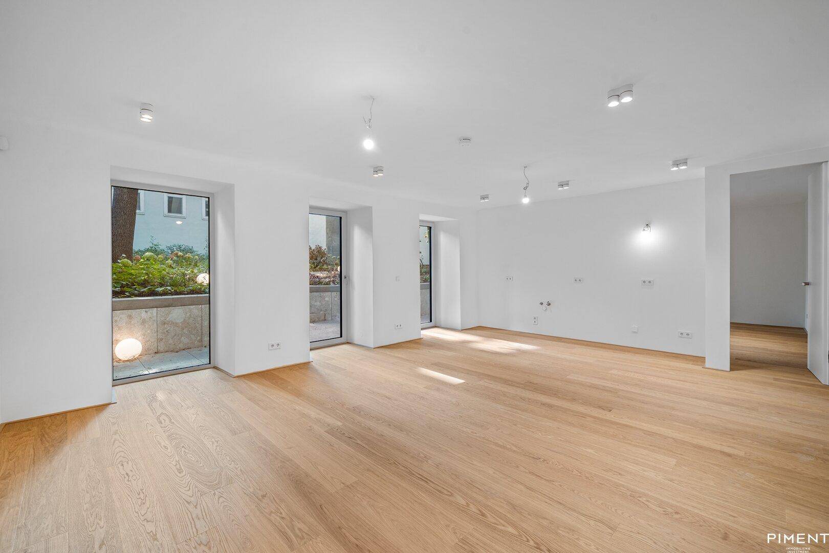 Heller, leerer Wohnraum mit Holzboden und großen Fenstertüren zum Gartenbereich.