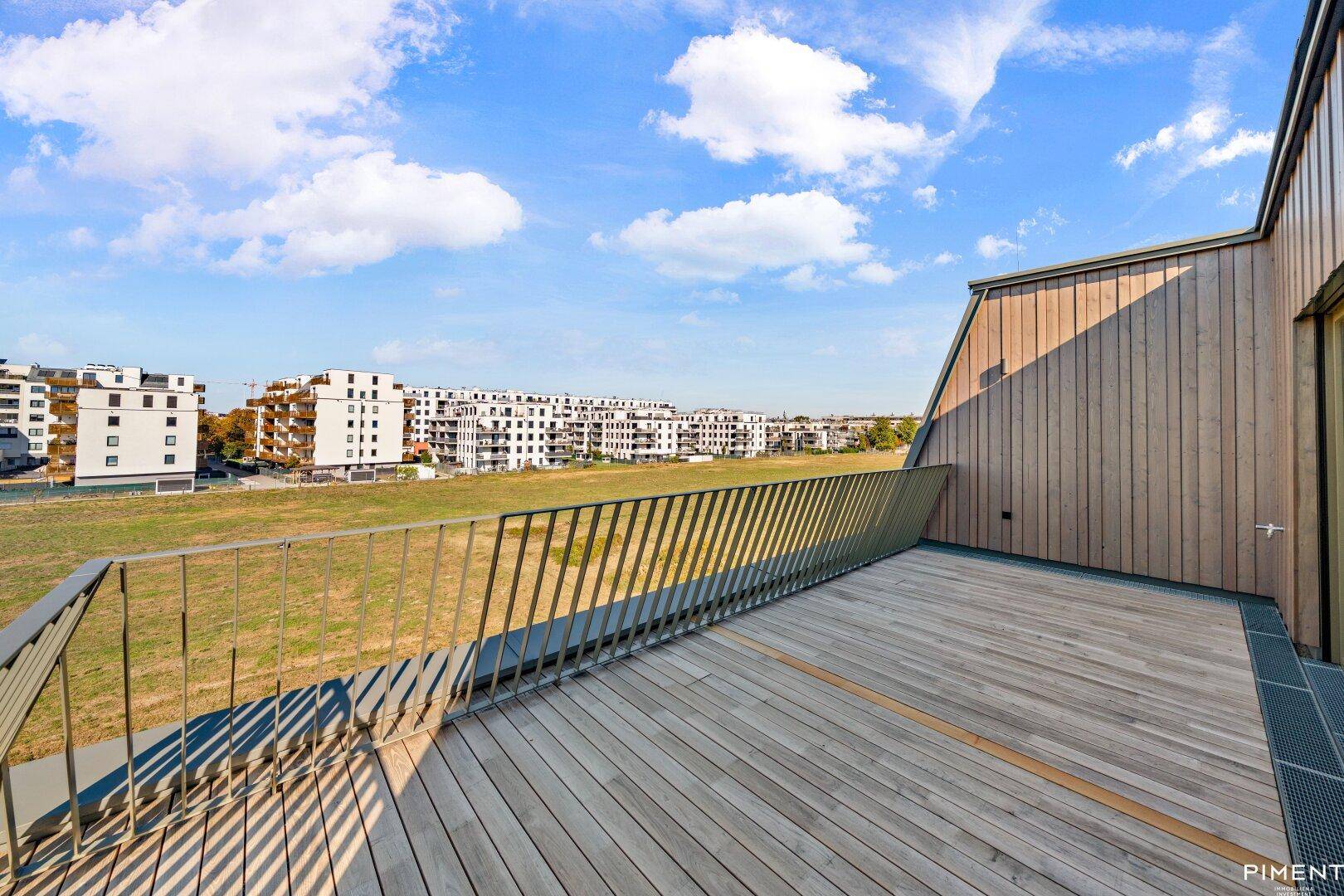 Große Holzterrasse mit Blick auf eine weite Grünfläche und moderne Wohngebäude in der Ferne.