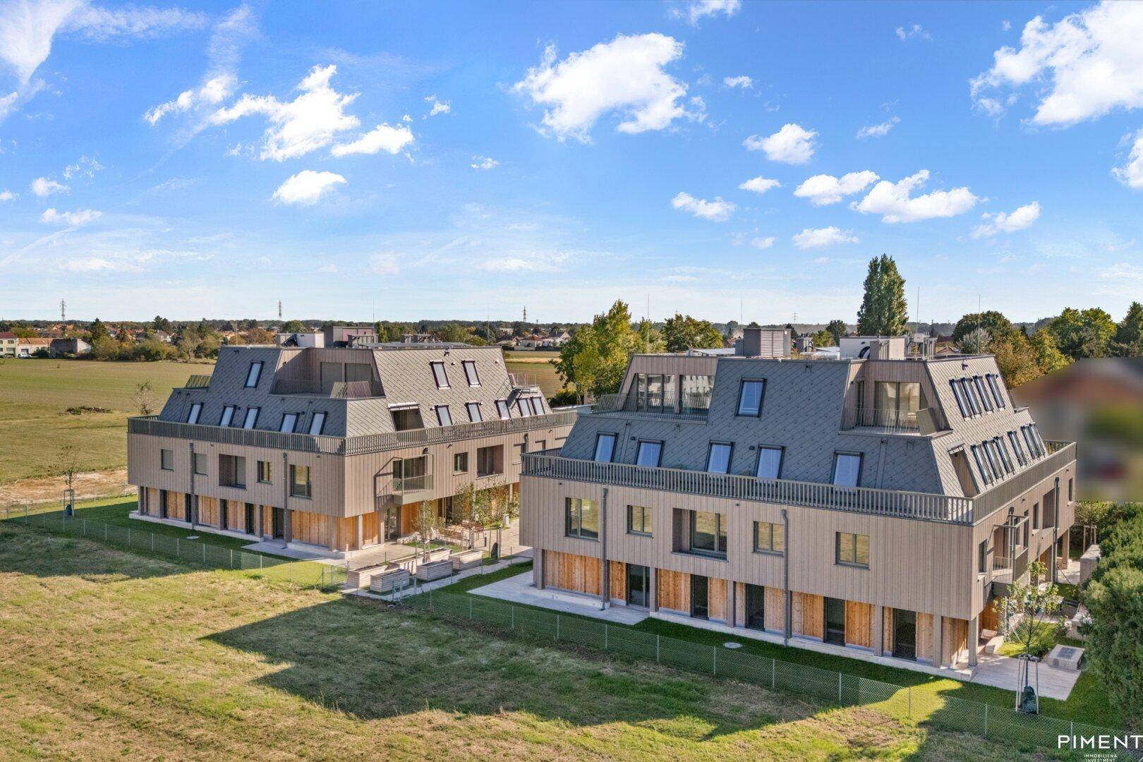Moderne Mehrfamilienhäuser mit Mansarddächern und Photovoltaikanlagen, umgeben von grüner Landschaft.