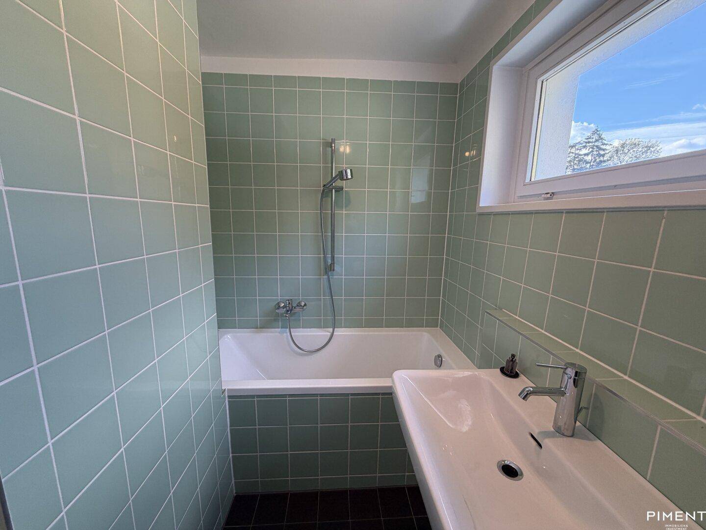 Badezimmer mit hellgrünen Fliesen, Badewanne mit Duschfunktion und einem modernen Waschbecken.
