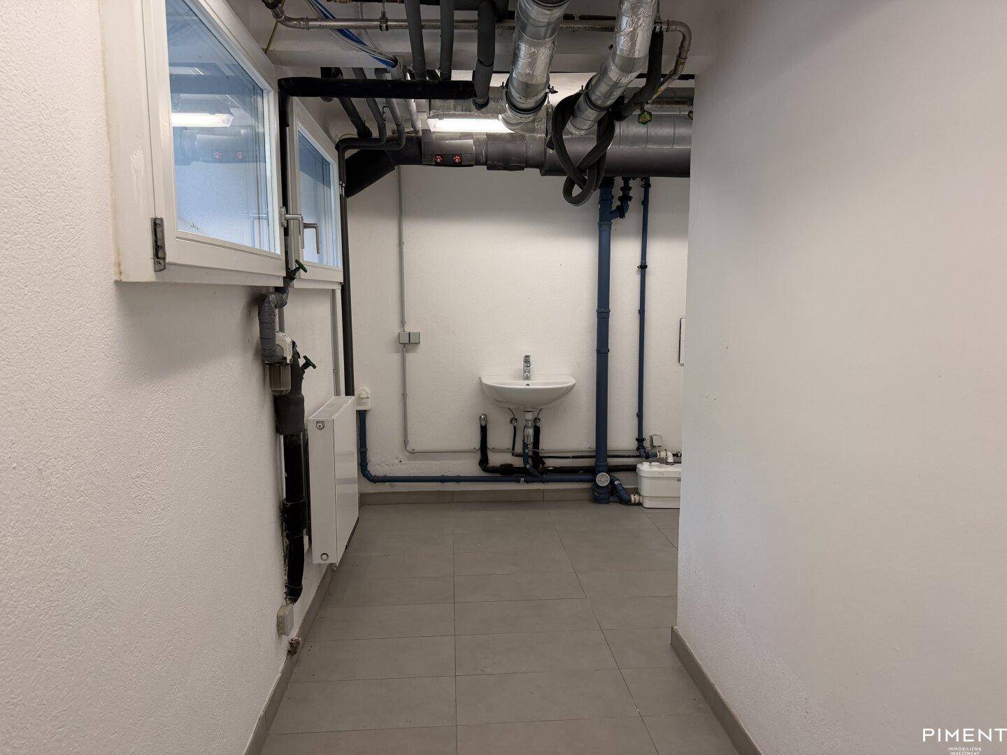 Technikraum im Keller mit sichtbaren Rohren, kleinem Waschbecken und Fenster.