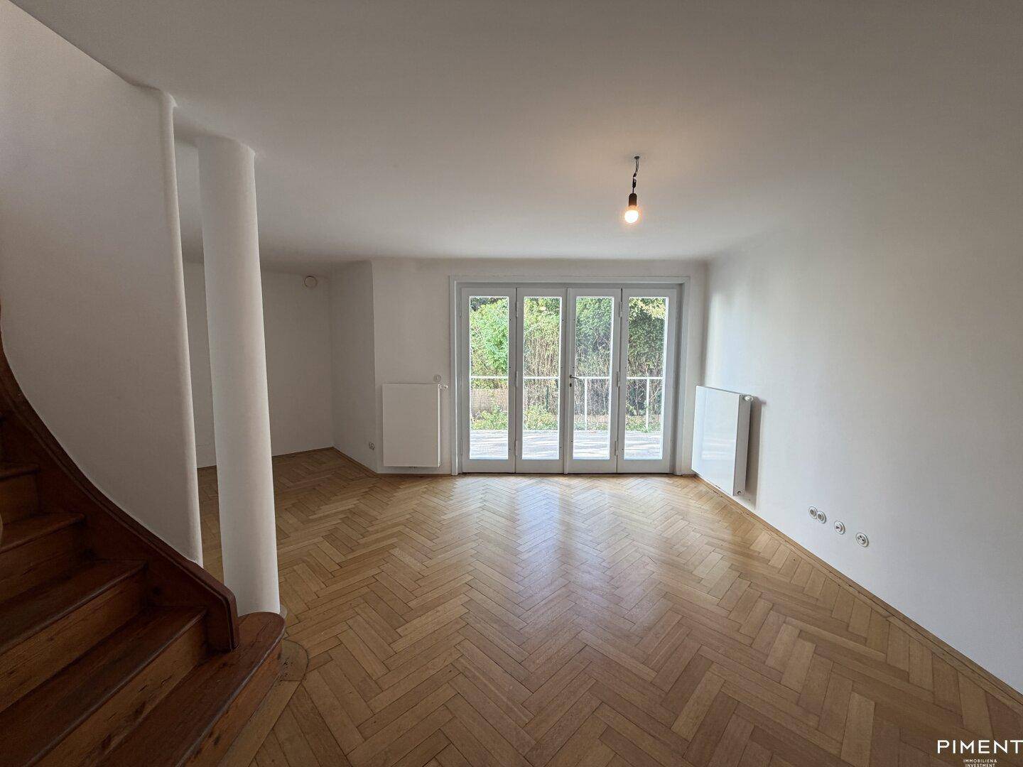 Helles Wohnzimmer mit Fischgrätparkettboden und großen Fenstertüren zum Außenbereich.