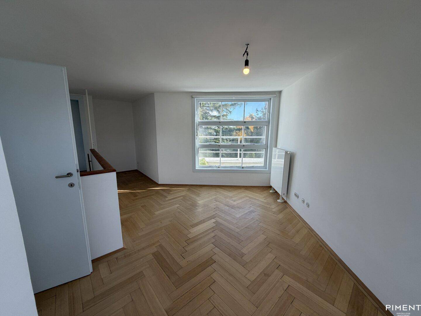 Geräumiges Zimmer mit Fischgrätparkett und großem Fenster, das viel Tageslicht hereinlässt.