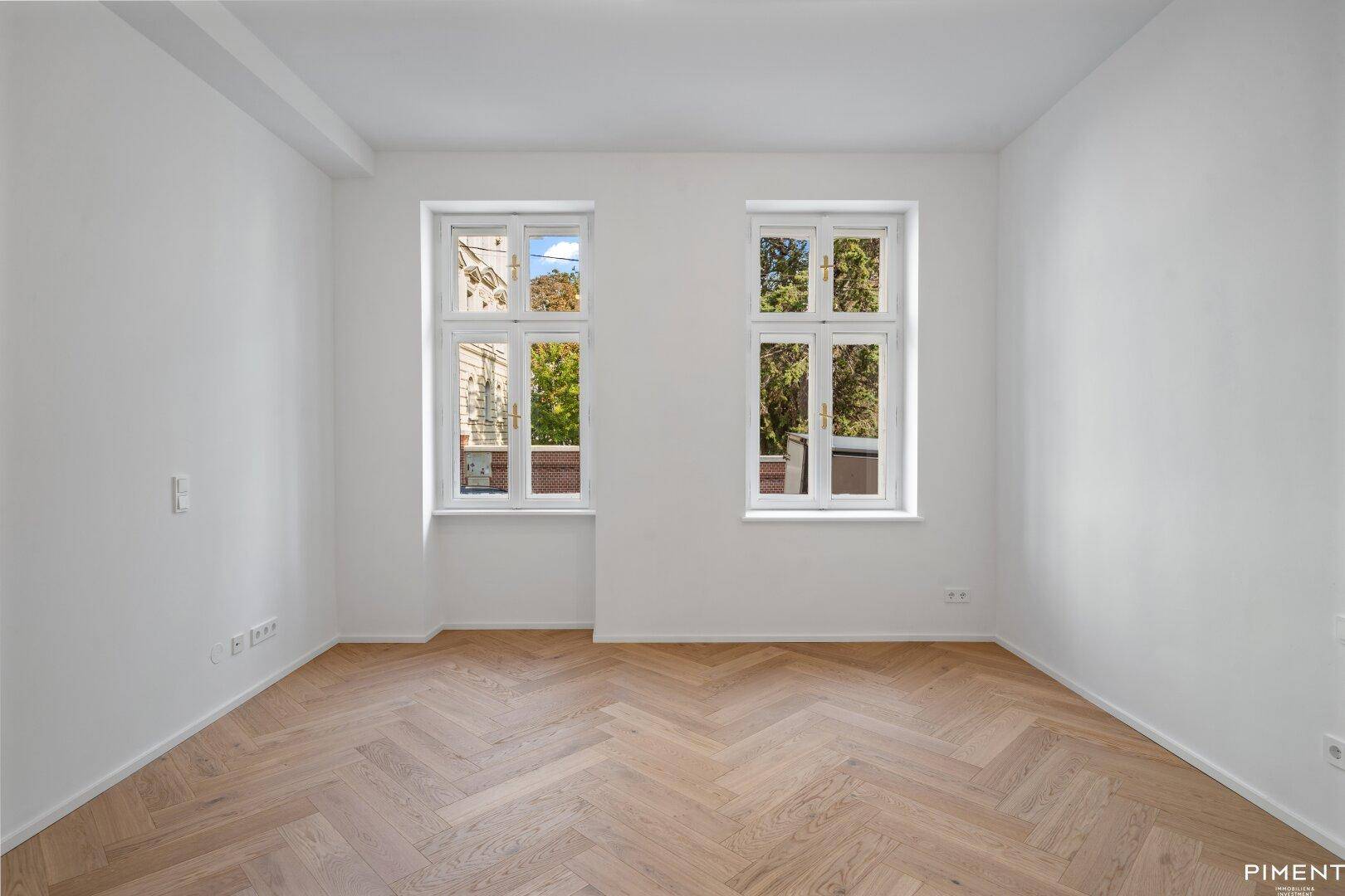 Lichtdurchflutetes Zimmer mit Fischgrätparkett und zwei hohen Fenstern, ideal für ein Schlafzimmer oder Büro.