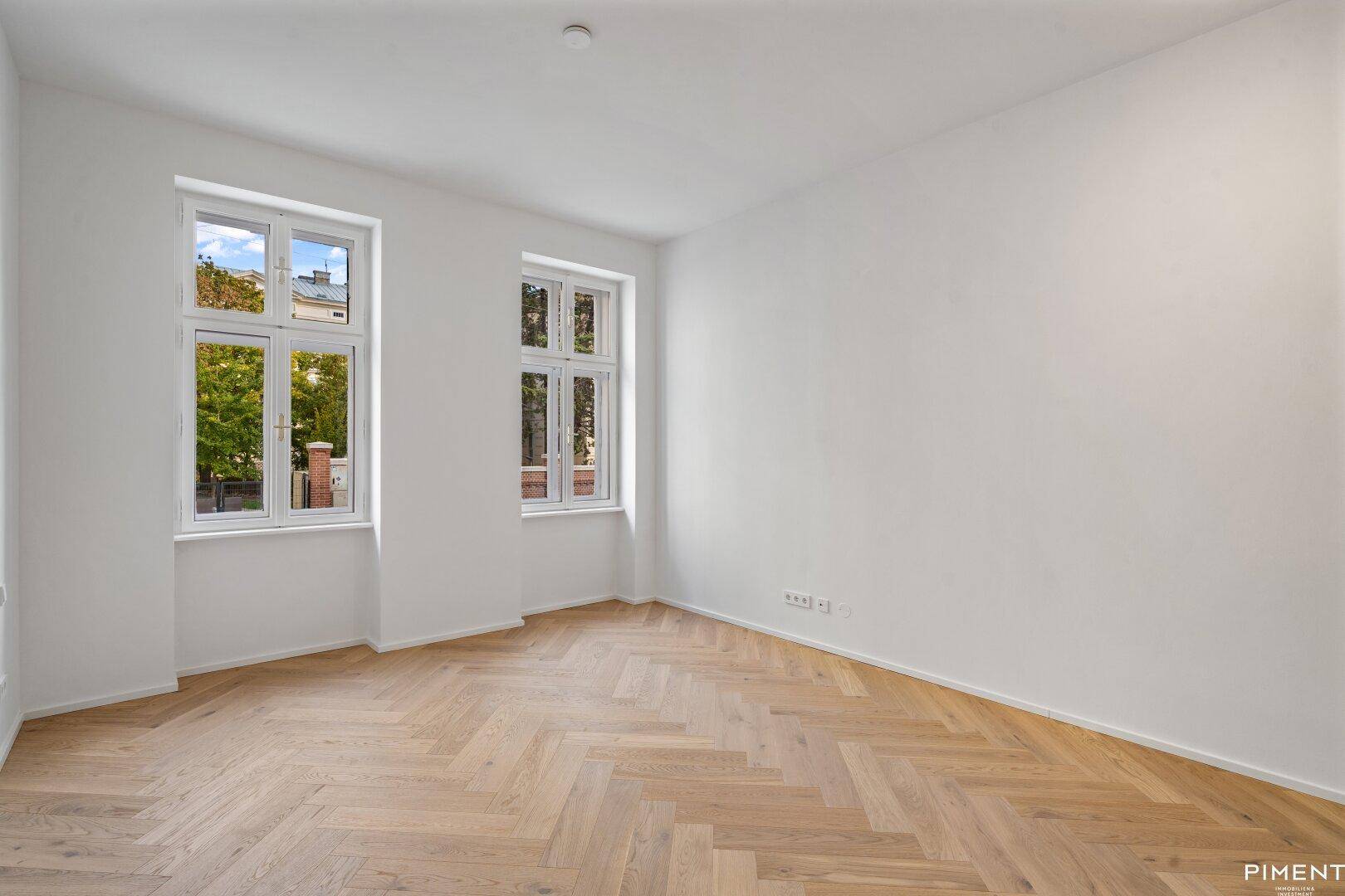 Geräumiges, helles Zimmer mit Fischgrätparkett und zwei großen Fenstern, ideal für Wohn- oder Schlafbereich.