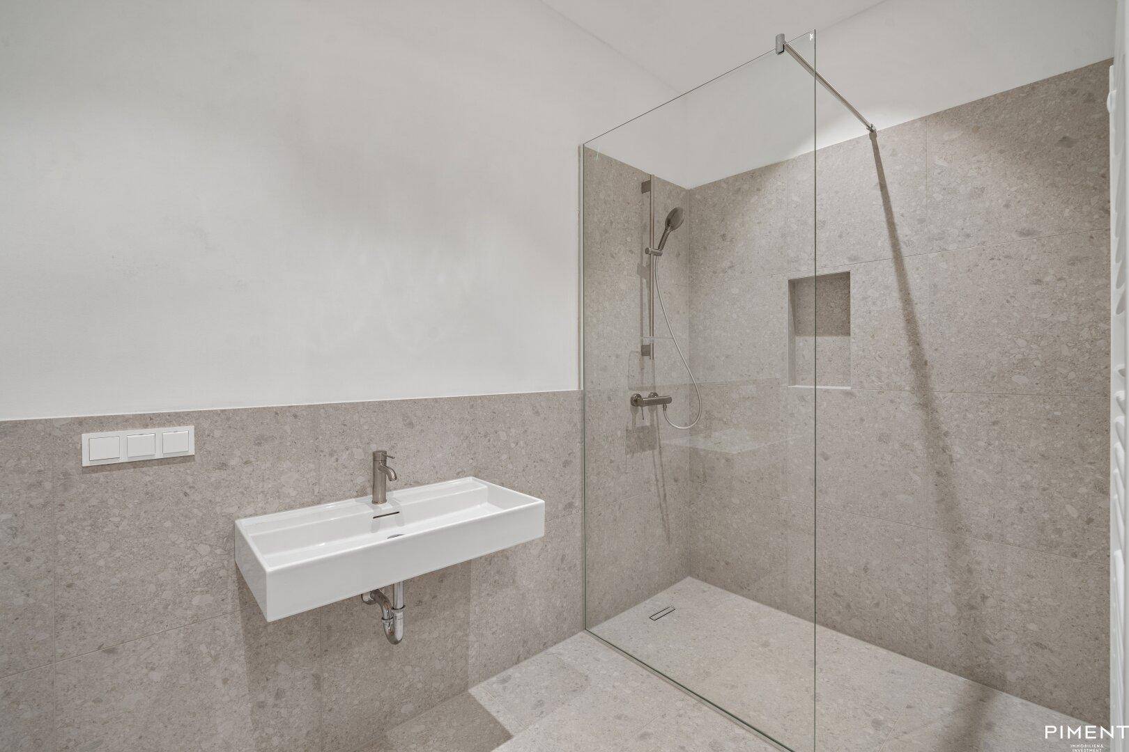Modernes Badezimmer mit großformatigen grauen Fliesen, ebenerdiger Dusche und Wandwaschbecken.