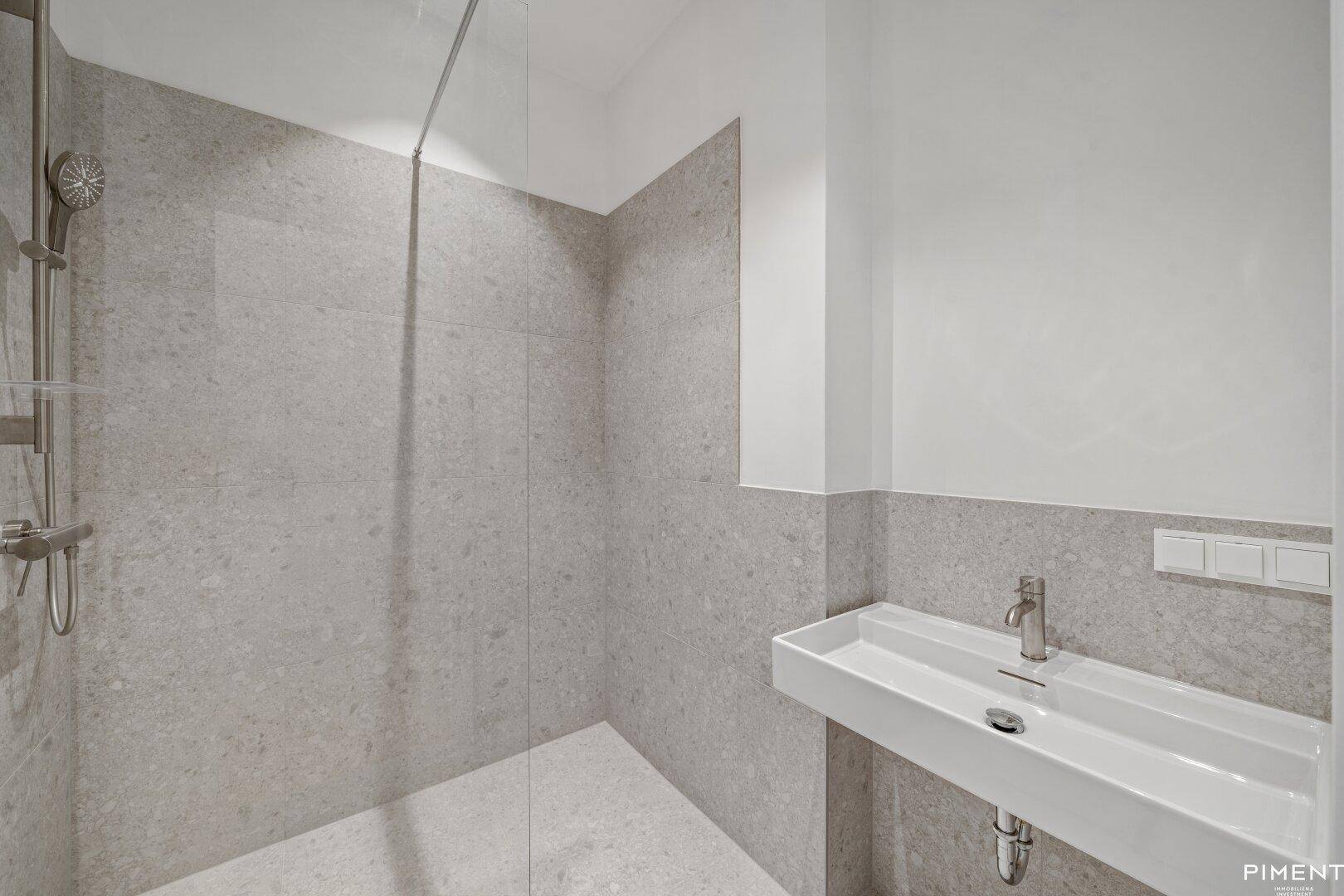 Modernes Badezimmer mit grauen Fliesen, einer Dusche und einem einzelnen Waschbecken.