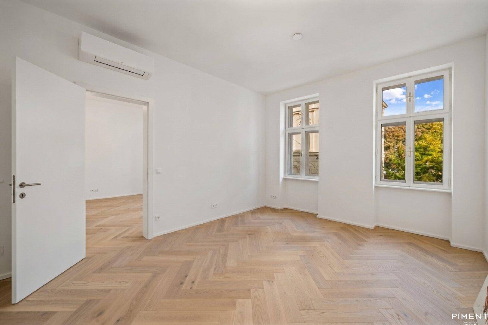 Heller Raum mit Fischgrätparkett, zwei Fenstern und einer Klimaanlage an der Wand.
