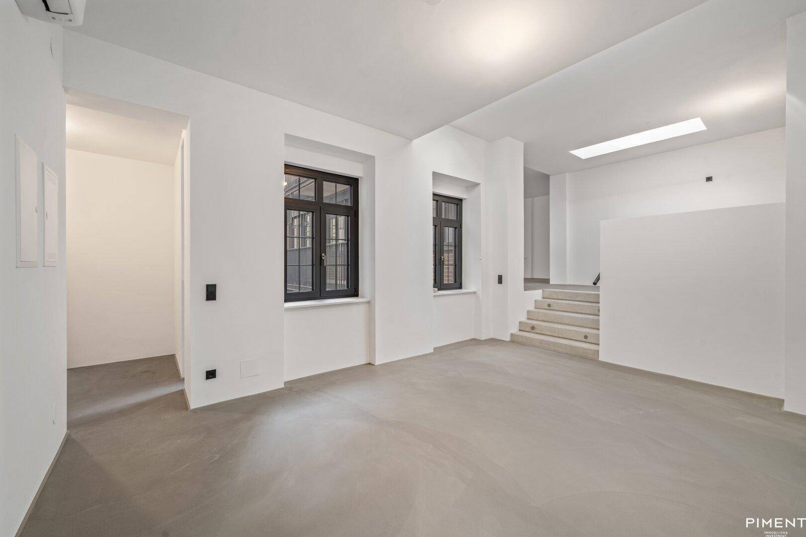 Geräumiger, unmöblierter Raum mit Betonboden, Oberlicht und Zugang zu einer Treppe.