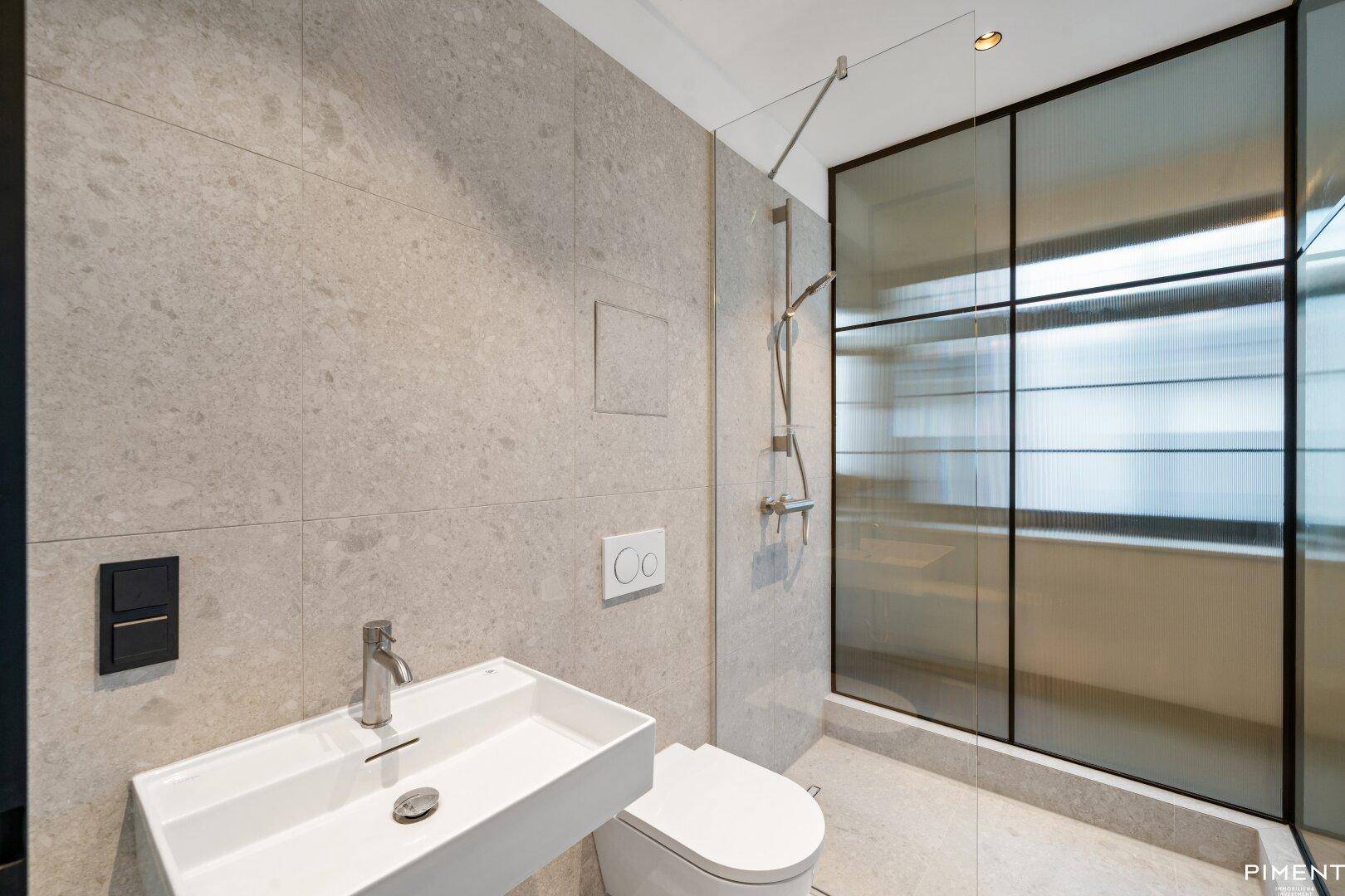 Modernes Badezimmer mit grauen Fliesen, wandhängendem WC und begehbarer Dusche.