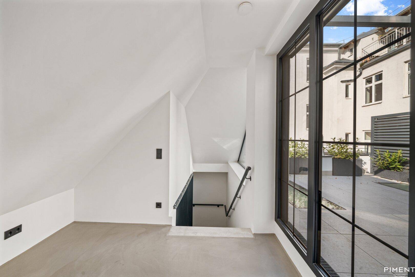 Dachgeschossraum mit schrägen Wänden, Betonboden und großem Fenster zur Terrasse.
