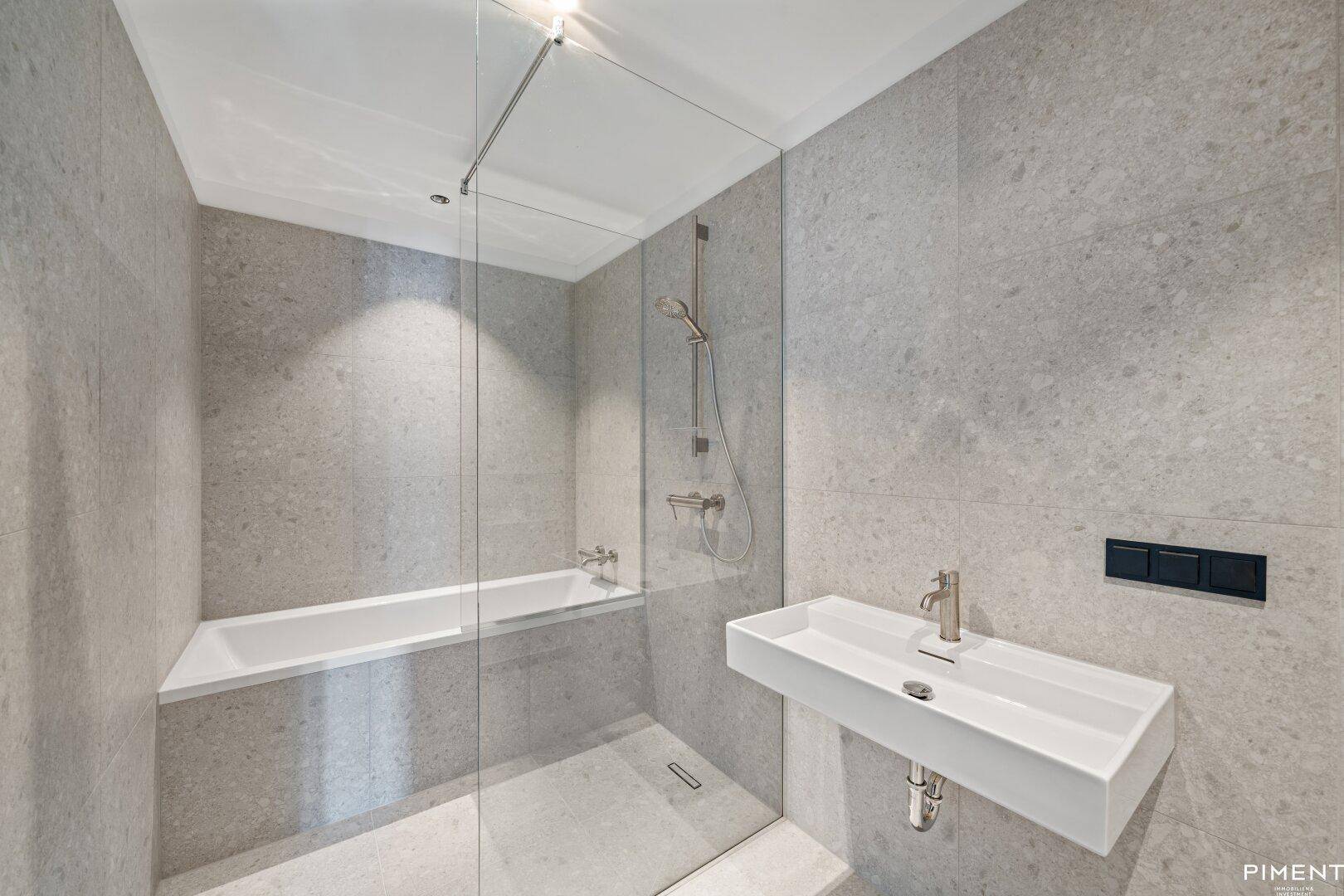 Modernes Badezimmer mit grauen Fliesen, Badewanne, Dusche und wandhängendem Waschbecken.