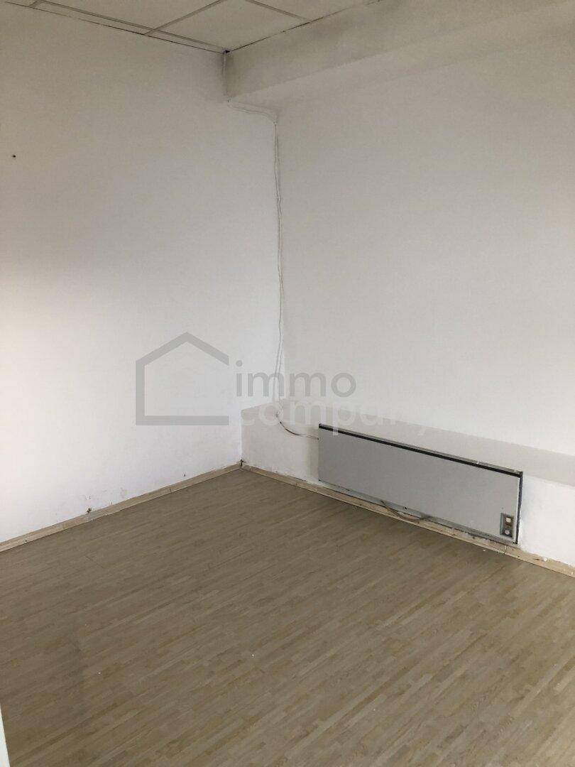 Leerer Büroraum mit weißen Wänden, Laminatboden, Heizkörper und abgehängter Decke.