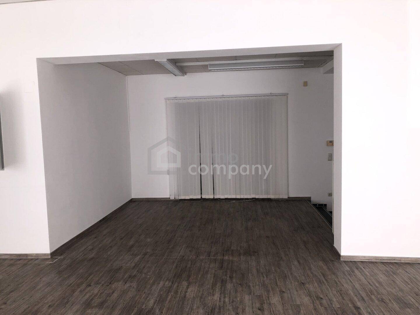 Heller, leerer Raumabschnitt mit vertikalen Jalousien vor dem Fenster und dunklem Bodenbelag.