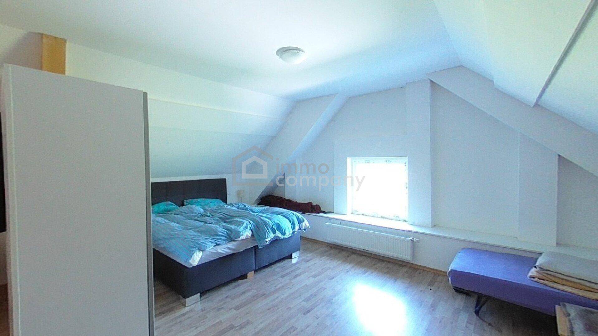 Helles Schlafzimmer im Dachgeschoss mit schrägen Wänden, Laminatboden und einem großen Fenster.