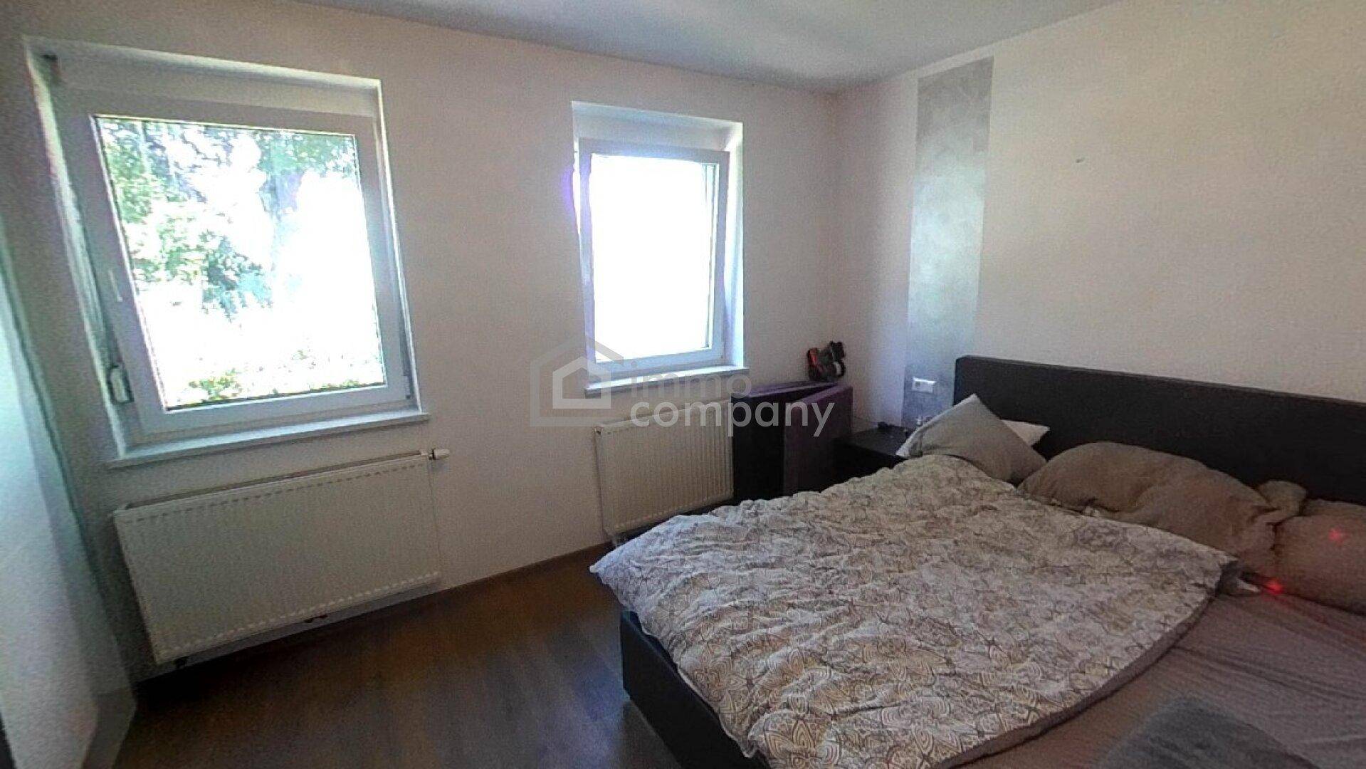 Schlafzimmer mit Doppelbett, zwei großen Fenstern und hellem Laminatboden.