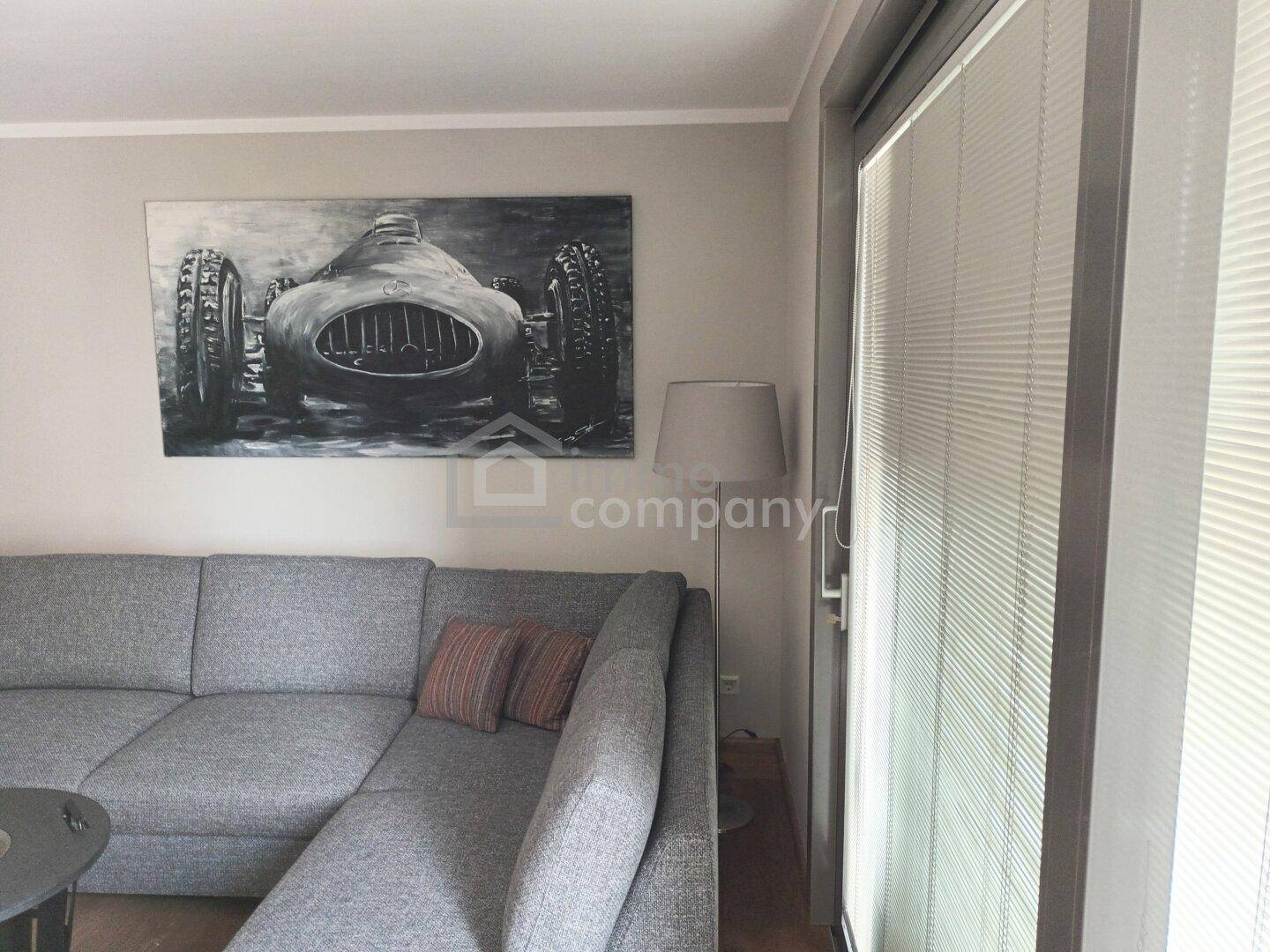 Gemütliches Wohnzimmer mit grauem Ecksofa, Kunstwerk an der Wand und großem Fenster mit Jalousien.