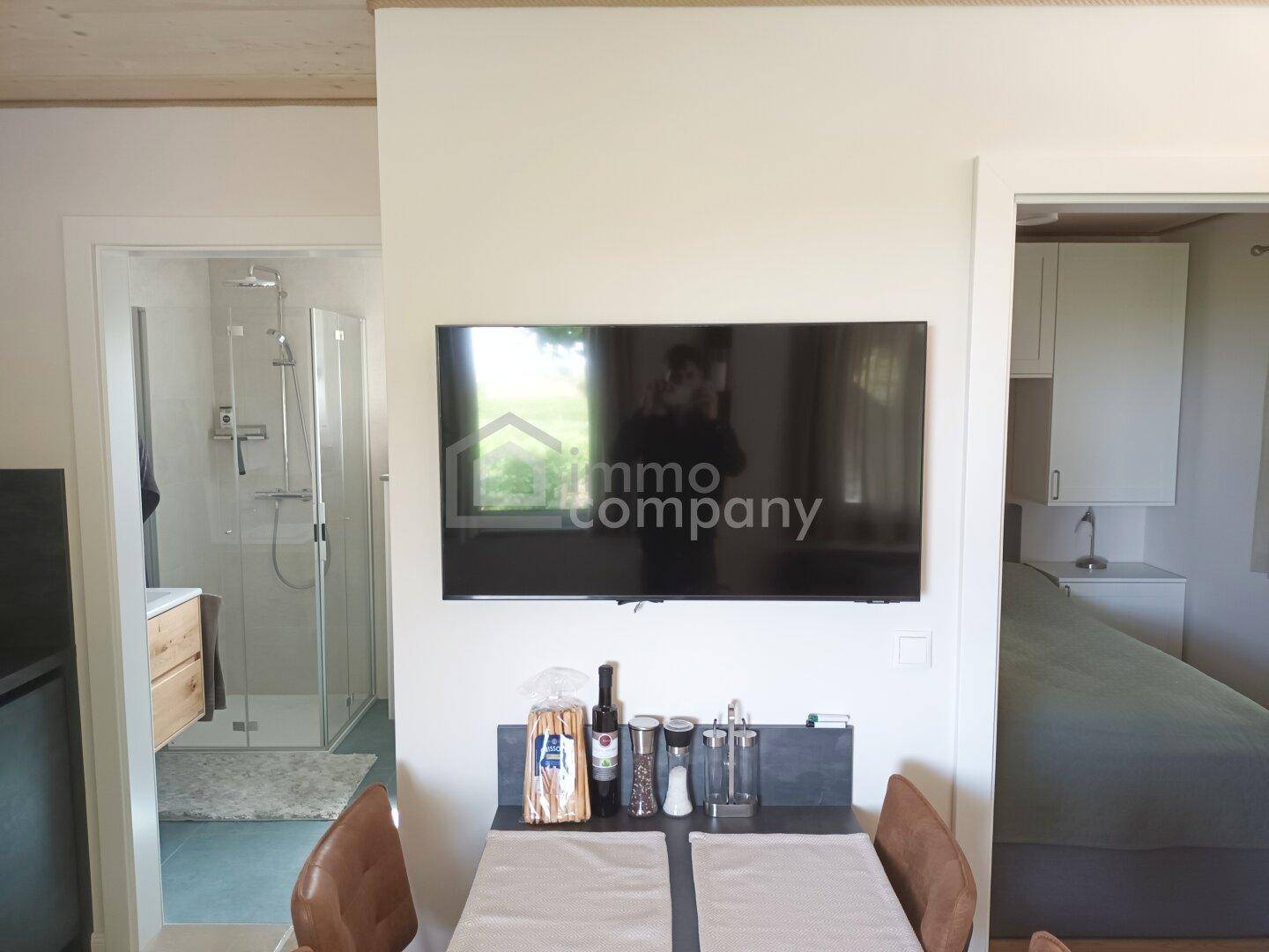 Innenraum mit Wandfernseher, Esstisch und Einblicken in ein Badezimmer und Schlafzimmer.