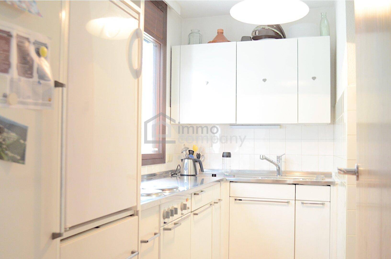 Funktionale Küche mit weißen Schränken, Edelstahlspüle und Fenster für Tageslicht.