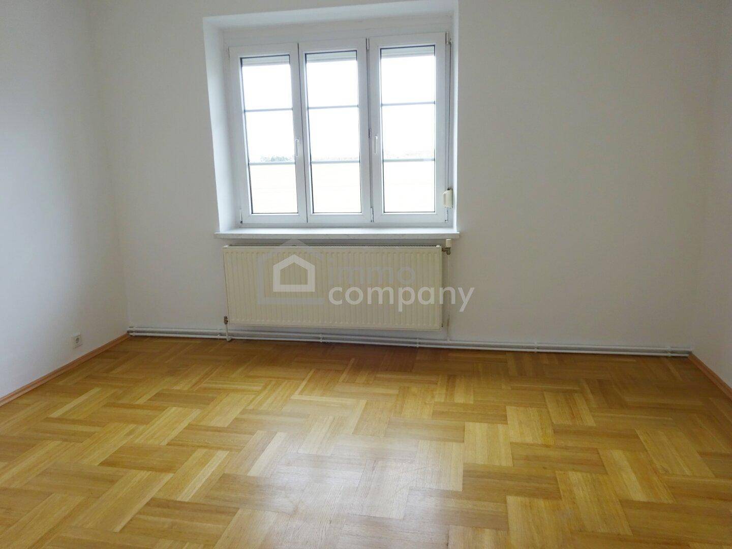 Leeres Zimmer mit Fischgrätparkettboden, weißen Wänden und einem großen Fenster.
