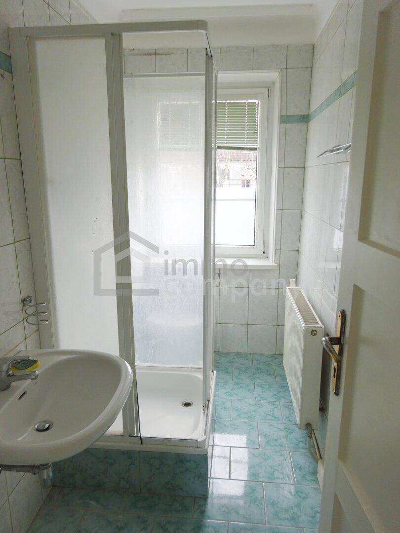 Badezimmer mit Duschkabine, Waschbecken, Heizkörper und Fenster, gefliest in Grün- und Weißtönen.