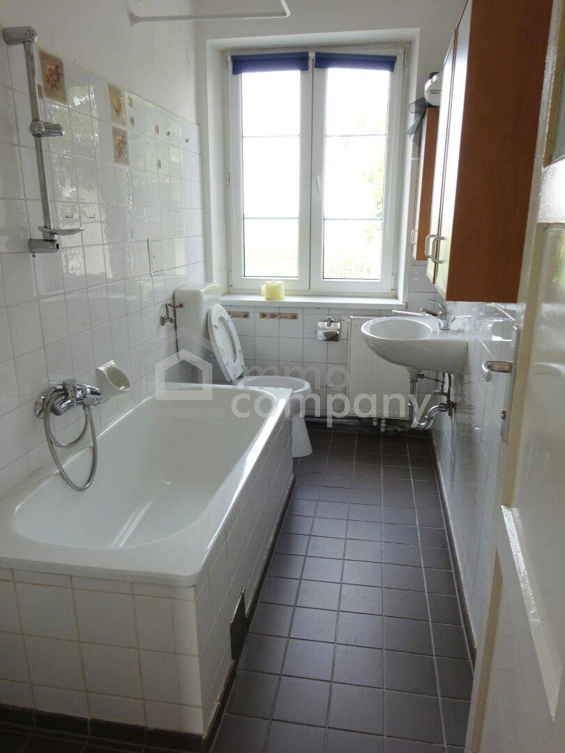Badezimmer mit Badewanne, WC, Waschbecken und Fenster, ausgestattet mit weißen Wandfliesen und dunklem Boden.