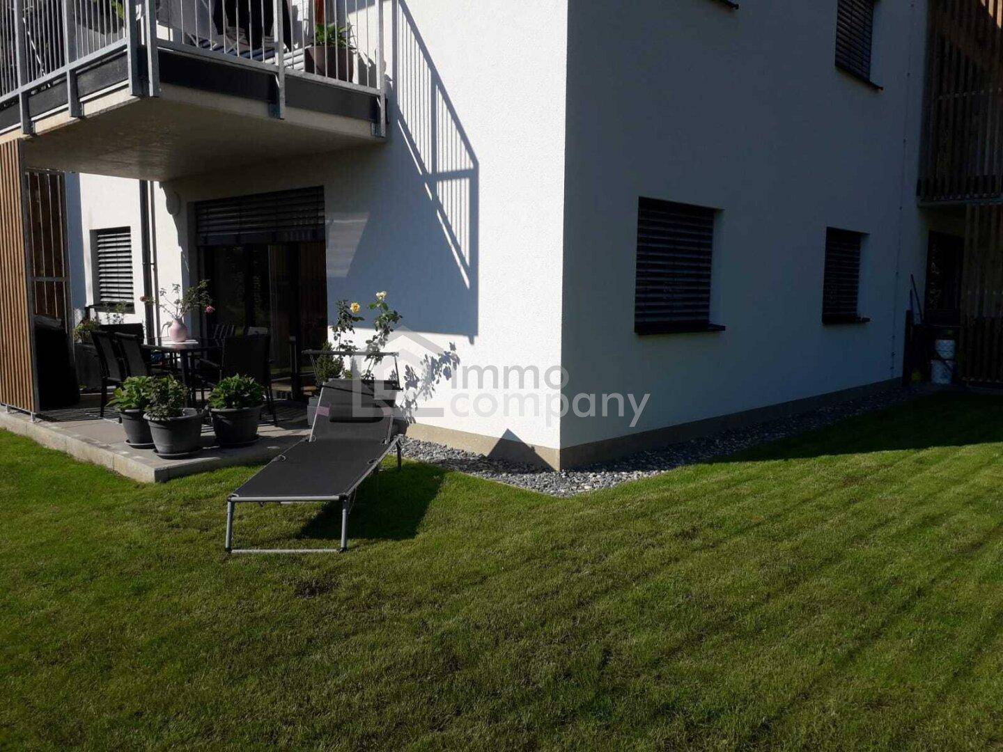 Blick auf die Terrasse und den Gartenbereich eines modernen Gebäudes an einem sonnigen Tag.