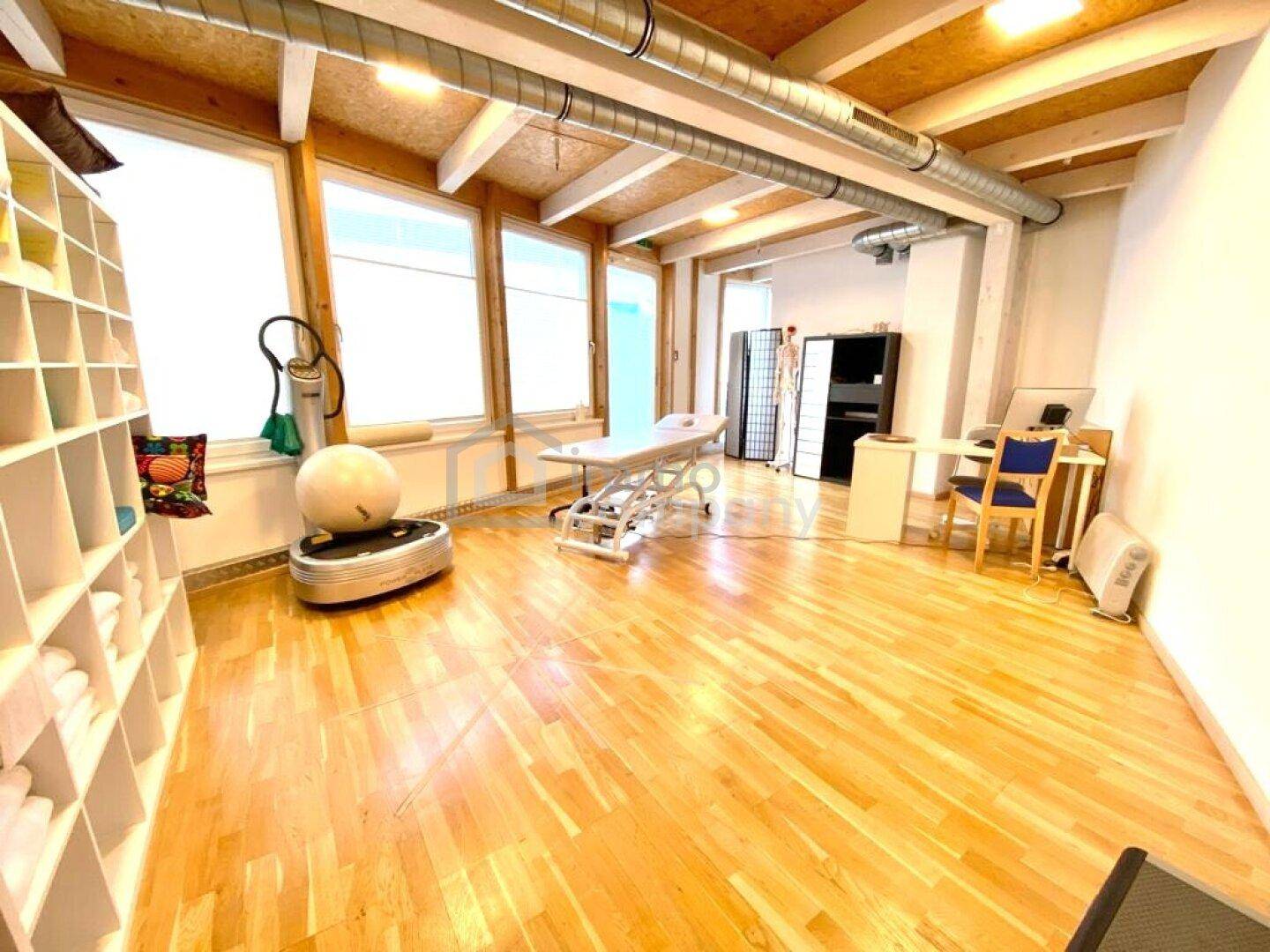 Geräumiger Therapieraum mit Holzboden, großen Fenstern und sichtbaren Deckenbalken, ausgestattet für Behandlungen.