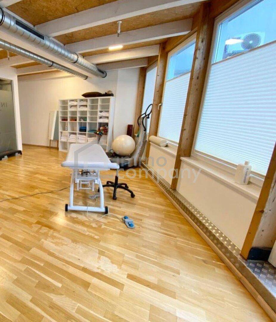 Blick in den Therapieraum mit Massageliege, Regalen und großen Fenstern, die viel Tageslicht hereinlassen.