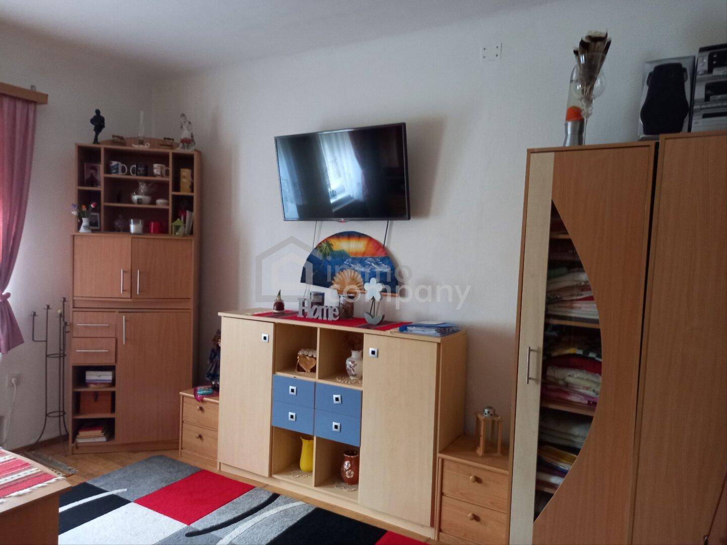 Gemütliches Wohnzimmer mit Fernseher, Holzmöbeln und einem farbenfrohen Teppich.