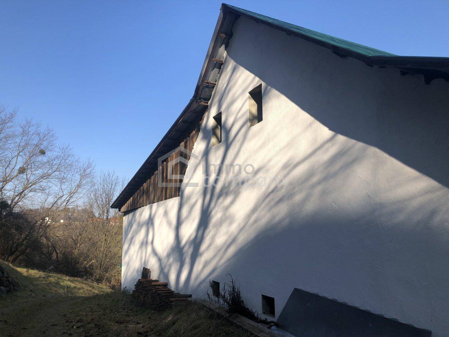Seitenansicht eines Gebäudes mit weißer Fassade, grünem Dach und Schatten von Bäumen.