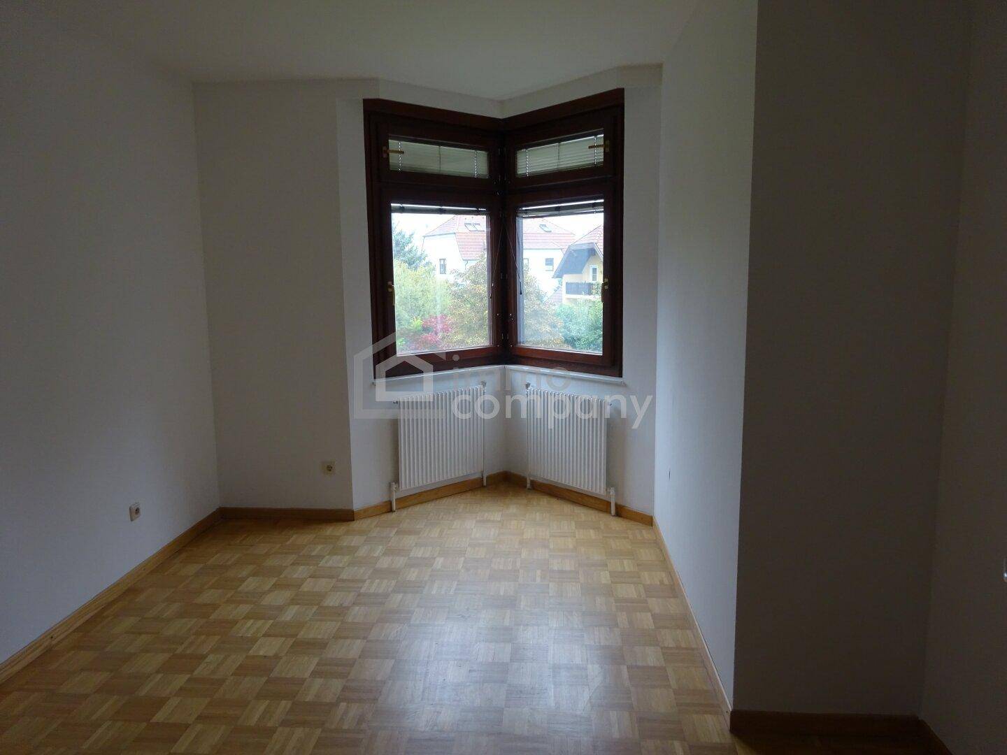 Heller Raum mit Parkettboden, weißen Wänden und einem Eckfenster mit Blick ins Grüne.