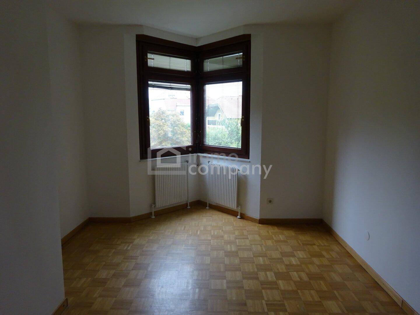 Geräumiger Raum mit Parkettboden, weißen Wänden und einem großen Eckfenster.