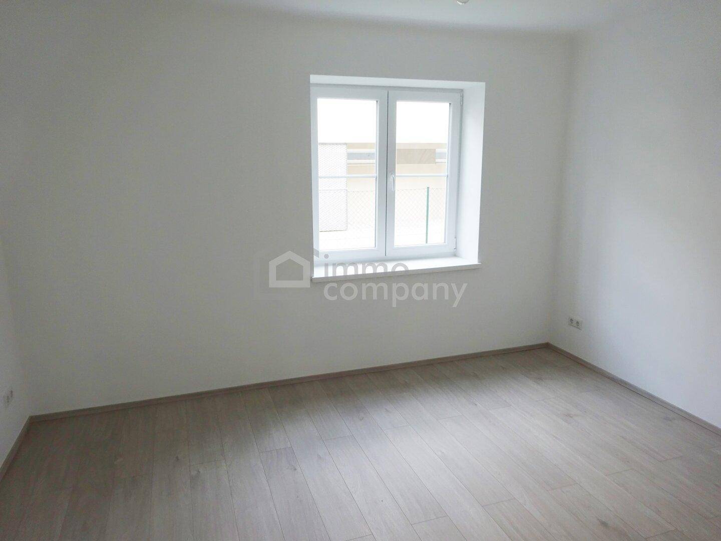 Helles, leeres Zimmer mit Laminatboden und Fenster, das viel Tageslicht hereinlässt.
