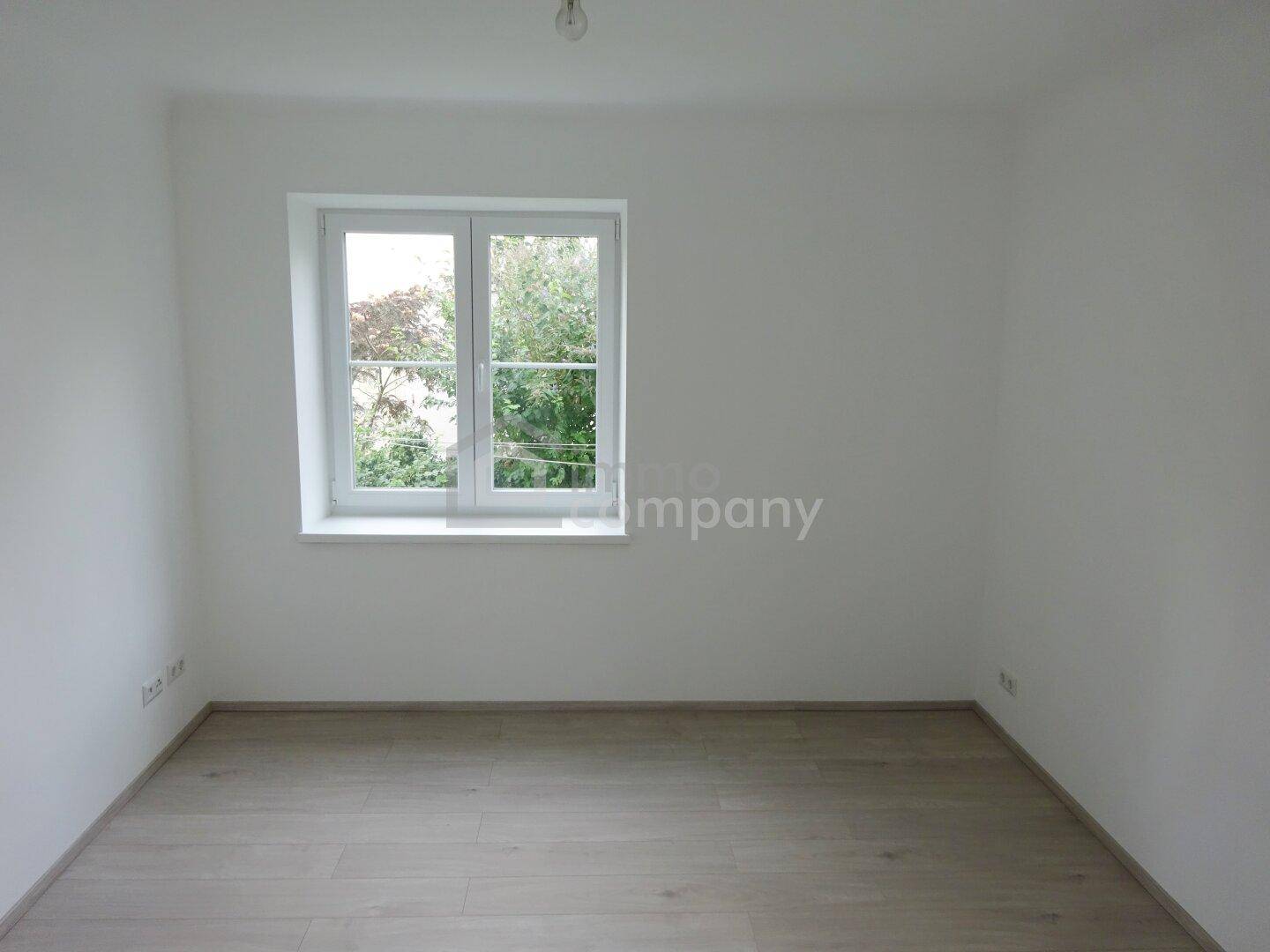 Leeres Zimmer mit hellem Laminatboden und einem Fenster mit Blick ins Grüne.