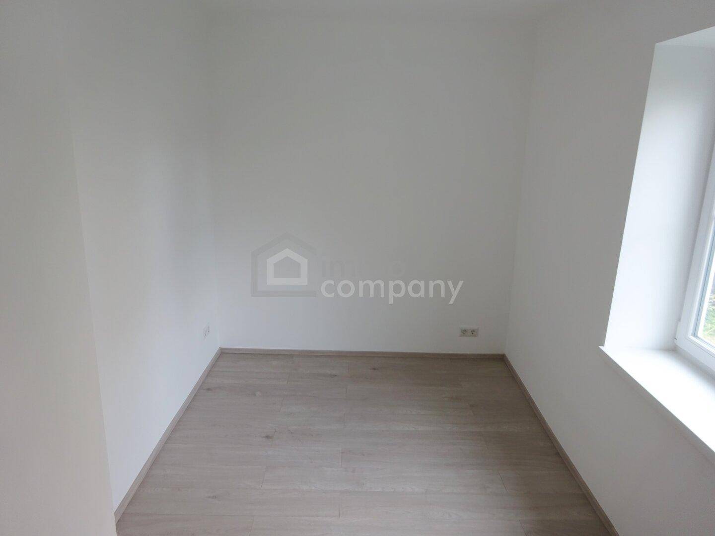 Leeres Zimmer mit hellem Laminatboden und einem Fenster, das seitliches Licht spendet.