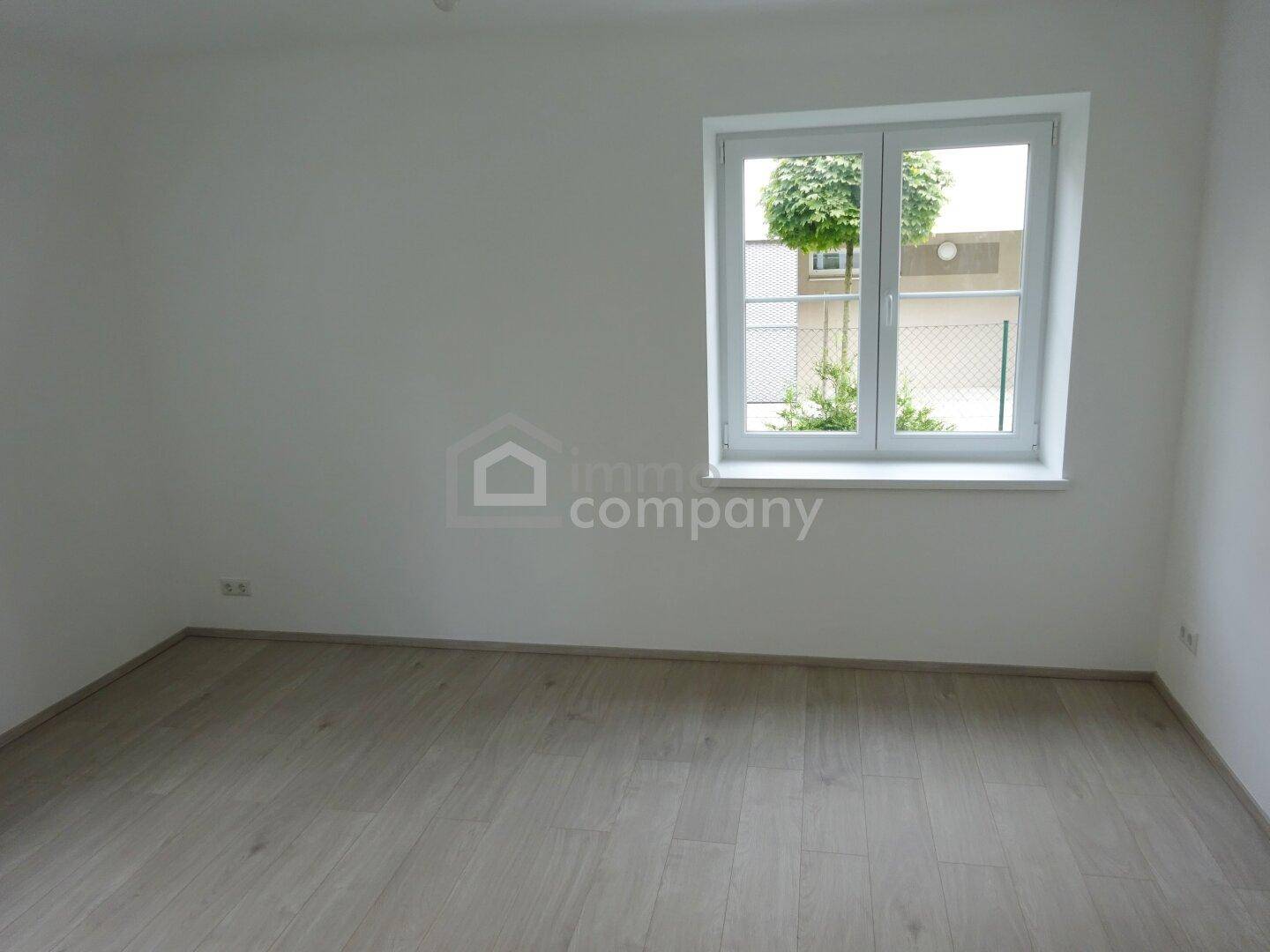 Helles, leeres Zimmer mit Laminatboden und Fenster, das einen Blick nach draußen bietet.