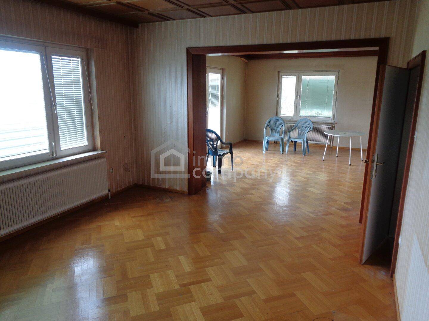 Geräumiges Wohnzimmer mit Parkettboden, Tapeten und Durchgang zu einem weiteren Raum.
