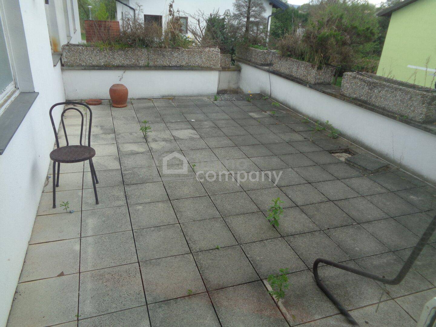 Große geflieste Terrasse mit Pflanzkübeln und einem Stuhl, renovierungsbedürftig.