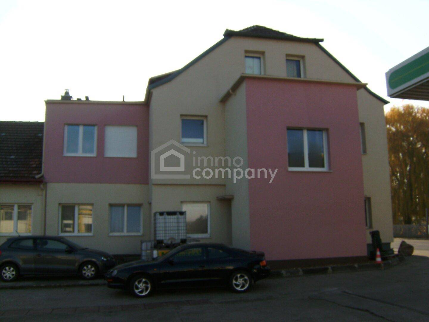 Seitenansicht eines Gebäudes mit rosa und beiger Fassade, mehreren Fenstern und zwei geparkten Autos davor.