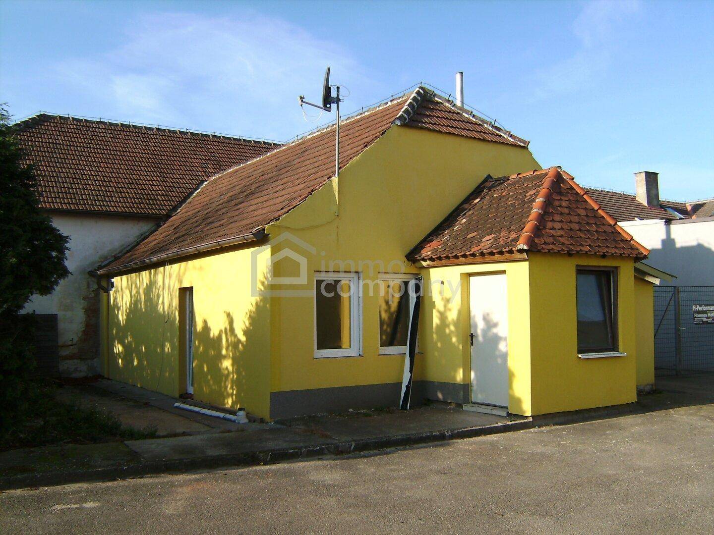 Gelbes Haus mit rotem Ziegeldach und einem kleinen Anbau, umgeben von einer gepflasterten Fläche.